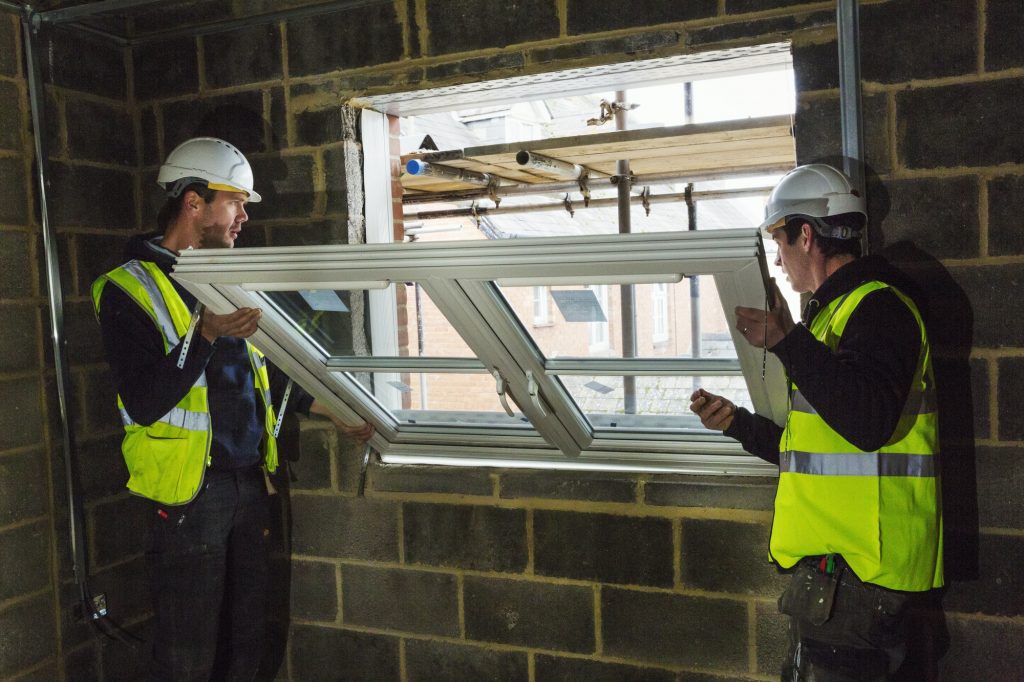 Two builders in hard hats and high visibility jacket fitting a uPVC window frame in Solihull.